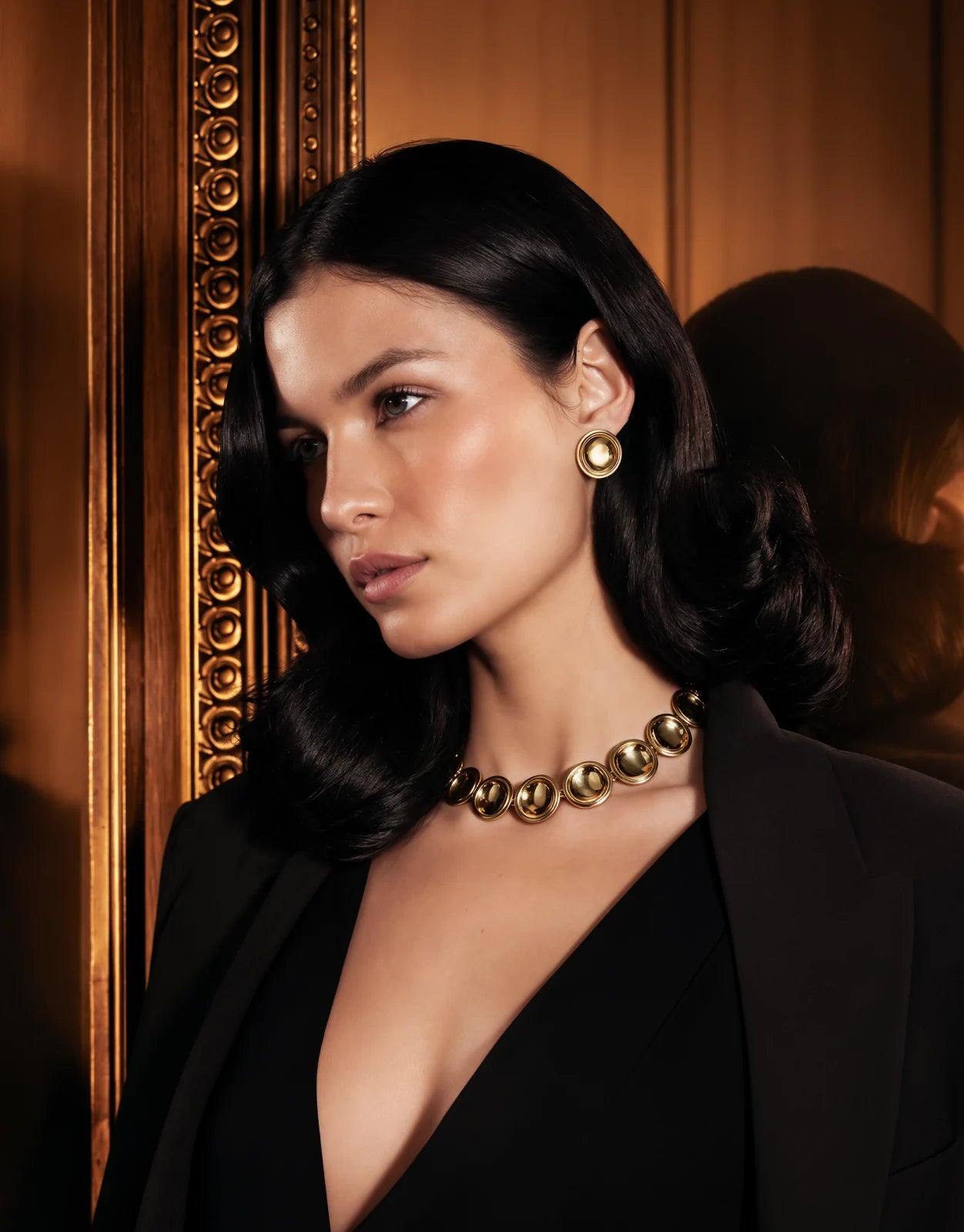 A model in a black jacket wearing the Patera Necklace and Earrings in a wood-panelled room