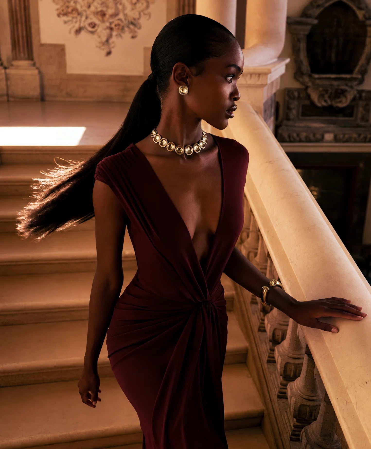 A model in a wine gown on a stone staircase, wearing the Patera Necklace, Earrings and Cuff