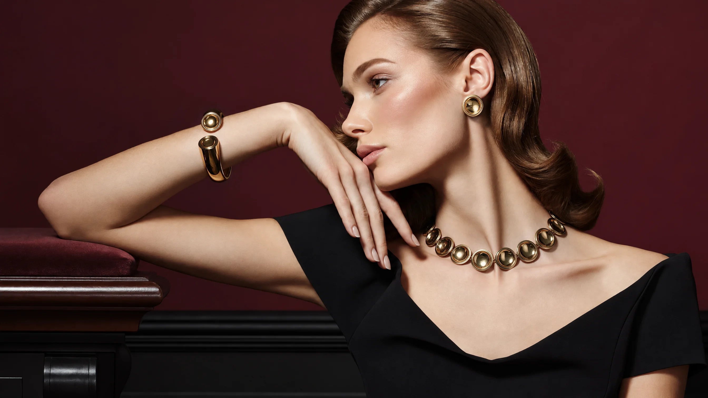 A model in a black dress leaning on one arm, wearing the Patera Necklace, Earrings and Cuff in gold