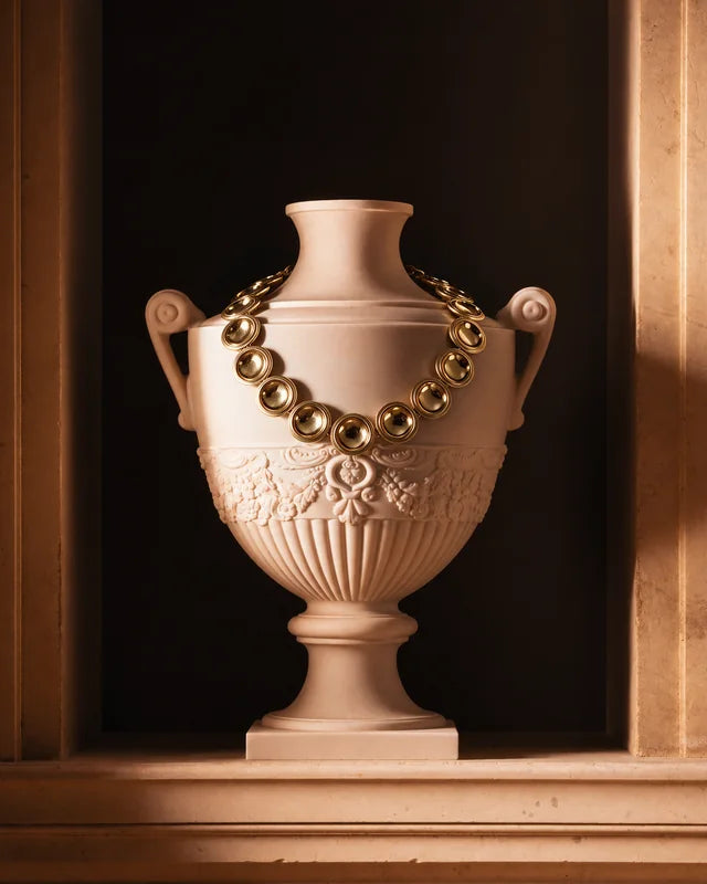 The Patera Necklace in gold draped over a carved stone urn